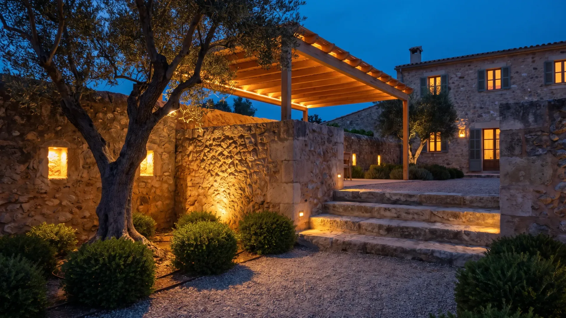 A Mallorcan garden terrace at dusk with an uplit olive, a grazed stone wall and recessed step lighting
